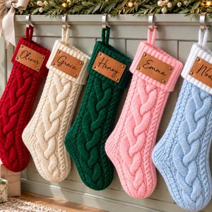 May include: Five cable-knit Christmas stockings in red, cream, dark green, light pink, and light blue. Each stocking features a white cuff with a rectangular brown leather patch engraved with the names Oliver, Grace, Henry, Emma, and Noah.