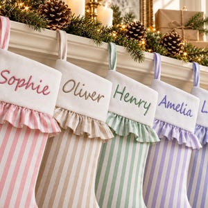 Könnte beinhalten: Fünf personalisierte Weihnachtsstrümpfe hängen in einer Reihe, jeder mit einer weißen Manschette und einem in passender Farbe gestickten Namen. Die Strümpfe haben vertikale Streifen in Rosa, Beige, Grün, Lavendel und Blau sowie einen Rüschenbesatz.