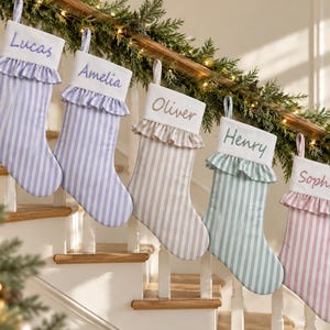 Puede incluir: Cinco calcetines navideños de tela con rayas verticales en morado claro, beige, verde y rosa. Cada calcetín tiene un puño blanco con borde de volantes y está bordado con los nombres Lucas, Amelia, Oliver, Henry y Sophie.