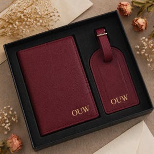 Puede incluir: Un portapasaporte y una etiqueta de equipaje a juego de color burdeos, presentados en una caja de regalo negra. Ambos artículos tienen un acabado texturizado y las letras OUW grabadas en oro. El portapasaporte es rectangular y la etiqueta tiene una correa.