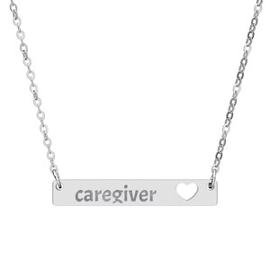 以下が含まれることがあります： 「caregiver」の文字とハート型の切り抜きが入った、シルバーのバーペンダントネックレス。繊細なチェーンが付いています。思いやりのあるジュエリーです。
