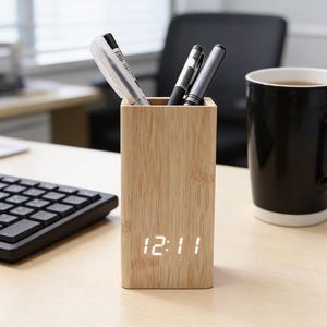 Pode incluir: Um porta-canetas retangular de bambu com um relógio digital que mostra 12:11. O suporte contém canetas e está ao lado de uma caneca preta. Há também um teclado na mesa.