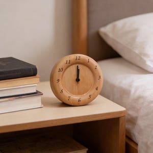Pode incluir: Um relógio redondo de madeira clara. O mostrador tem algarismos e ponteiros pretos, indicando que são pouco depois das 12 horas. O relógio está numa mesa de cabeceira clara, ao lado de uma cama com lençóis brancos.