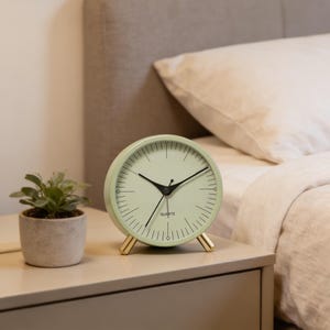 May include: A mint green quartz clock with black hands and gold-colored legs sits on a nightstand. The clock face has black markings and the word "QUARTZ" at the bottom. A small potted plant is to the left of the clock.