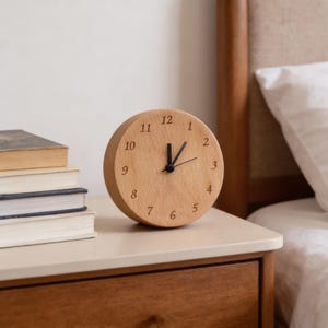 May include: A round wooden clock with black hands and numerals. The clock sits on a nightstand next to a stack of books. The clock's face is light brown, and the numbers are etched into the wood. The clock is approximately 4 inches in diameter.