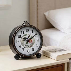 Op de afbeelding: Een zwart en bronskleurige analoge klok met een witte wijzerplaat met zwarte cijfers en een bloemenontwerp. De klok heeft een klein handvat aan de bovenkant en staat op twee zwarte bolvormige voetjes. Er ligt een boek naast de klok.