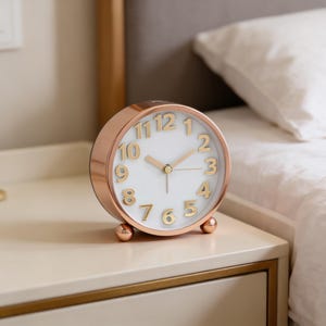 May include: A rose gold analog alarm clock with a white face and gold numerals. The clock has a round shape and sits on two small, round feet. The hands are gold, and the clock is on a white nightstand.