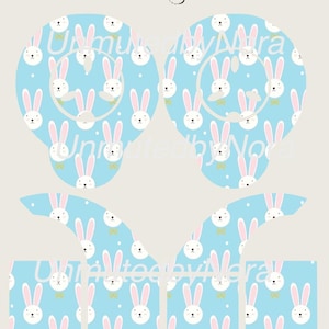 Puede incluir: Diseño azul claro con caras de conejos blancos, orejas rosas y lazos amarillos. El diseño se repite en varias formas, incluyendo dos grandes formas de orejas y formas rectangulares más pequeñas. También está presente el texto "Unmuted by Vera".