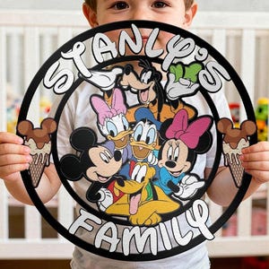 Könnte beinhalten: Ein rundes, schwarzes Wandschild mit dem weißen Schriftzug "STANLY'S FAMILY". In der Mitte befindet sich eine farbenfrohe Illustration von Mickey Maus, Minnie Maus, Donald Duck, Daisy Duck, Goofy und Pluto, flankiert von zwei Eiswaffeln.