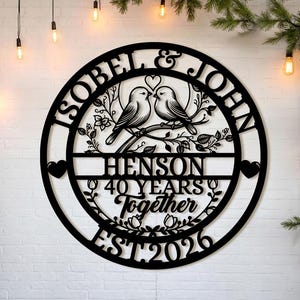 May include: A black metal wall art piece featuring a circular design with the names "Isobel & John" and "Henson". It includes two birds on a branch, the words "40 Years Together", and the year "EST 2026".