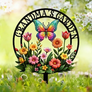 以下が含まれることがあります： 「GRANDMA'S GARDEN」の文字が円形にデザインされた黒い金属製のガーデンステーク。カラフルな蝶が円の中心に配置され、鮮やかな花々のベッドの上にあります。花には、チューリップ、デイジー、その他の花が含まれています。