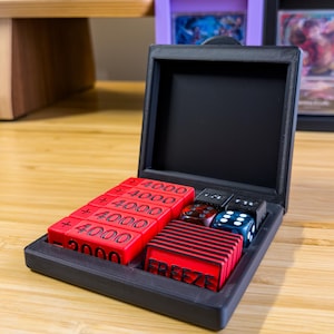 May include: A black rectangular storage case containing five red square tokens marked with +4000, one red token marked with -2000, a stack of red tokens labeled FREEZE, and four multi-colored six-sided dice nestled in dedicated compartments.
