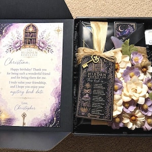 May include: A black gift box containing a greeting card with purple floral and nautical illustrations, a wrapped book with a gold ribbon, a decorative key tag labeled The Hidden Bookcase, paper flowers in shades of purple and cream, and a small digital timer.