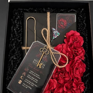 May include: A black gift box containing a black envelope, a large gold paperclip, a small decorative key tied with twine, and a cluster of red fabric roses. A clear card features the text: Hidden Book, This book is a game of secrets.