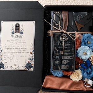 May include: A black gift box containing a book wrapped in black paper, a decorative metal key, a black card with text, a blue butterfly ornament, and assorted blue, orange, and cream fabric flowers. A letter with the text The Hidden Bookcase is attached.