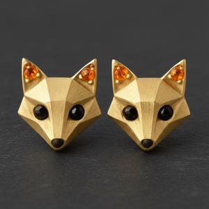 May include: A pair of fox-shaped stud earrings with a brushed gold finish. Each earring features a geometric, faceted design with two small, round, orange amber stones set in the ears and two round, black onyx stones serving as the eyes.