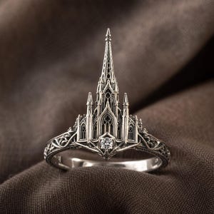 May include: A silver ring featuring a detailed cathedral design. The ring has a central spire and intricate architectural details, with a small, clear gemstone at the base of the spire. The band has an ornate, filigree pattern.