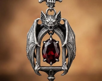 May include: A silver-toned metal pendant featuring a detailed bat with outstretched wings framing a central oval-shaped deep red gemstone. The top of the pendant is designed as a skeletal hand grasping a horizontal bone-shaped bail.