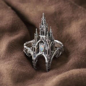 May include: A silver-colored ring featuring a detailed gothic cathedral design. The ring has a central oval-shaped opal stone and intricate architectural details, including spires and arched windows. The ring is set against a brown fabric background.
