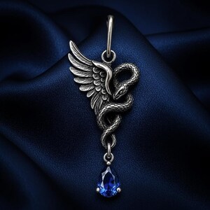 Peut inclure: Un pendentif en métal argenté représentant un serpent enroulé détaillé avec une aile emplumée s'étendant sur le côté. Le pendentif comprend un petit anneau de jonction en haut et une pierre précieuse bleue facettée en forme de goutte.