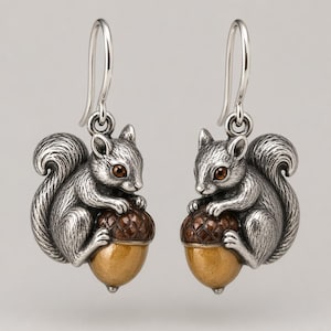 May include: A pair of silver-toned drop earrings featuring a detailed squirrel figure holding an acorn. The squirrel has a textured metallic finish with dark eyes, while the acorn has a brown textured cap and a smooth, light brown base.