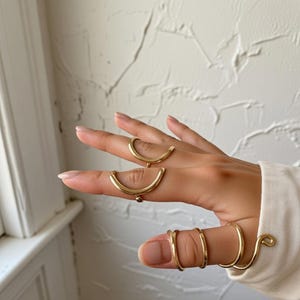 Puede incluir: Una mano con varios anillos dorados. Los anillos tienen diseños únicos, incluyendo formas curvas y en espiral. El fondo es una pared blanca texturizada, y la persona lleva una camisa blanca de manga larga.