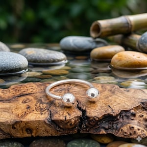 Op de afbeelding: Een zilverkleurige open manchetarmband met twee bolvormige uiteinden. De armband wordt getoond op een stuk hout met een natuurlijk nerfpatroon, met een vage achtergrond van water, stenen en groen.