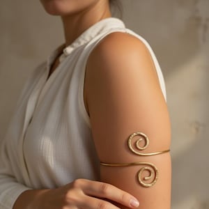 Pode incluir: Uma pulseira de braço dourada com um design em espiral. A pulseira é usada na parte superior do braço e apresenta dois detalhes em espiral martelados. A joia é um acessório simples, mas elegante.