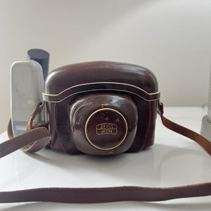 May include: A vintage brown leather camera case with a matching strap. The case has a rounded shape with white trim and a circular emblem that reads "ZEISS IKON". A silver flash unit is visible to the left.