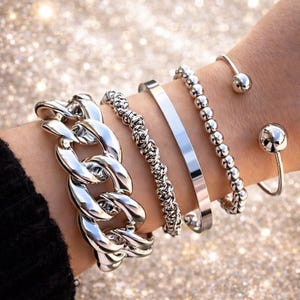 Puede incluir: Primer plano de una colección de pulseras plateadas en una muñeca. Las pulseras incluyen una cadena gruesa, un diseño de cuerda trenzada, una banda lisa, un estilo de cuentas y dos pulseras de puño abiertas con extremos de bola. El fondo es brillante.