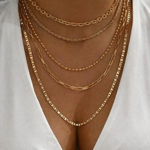 Könnte beinhalten: Eine Sammlung von geschichteten goldenen Halsketten. Die Halsketten weisen verschiedene Kettenstile auf, darunter Kabel-, Perlen- und Flachglied-Designs. Der Schmuck wird vor einem weißen Oberteil präsentiert.