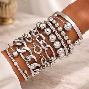 Può includere: Una collezione di braccialetti argentati adorna un polso, mostrando vari design. Questi includono maglie a catena spesse, fili di perline e sfere lisce e arrotondate. I braccialetti sono esposti su uno sfondo morbido e neutro, con il marchio "BARVEDA" visibile.