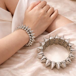 Może przedstawiać: Srebrne bransoletki z kolcami. Jedna bransoletka jest noszona na nadgarstku, a druga leży obok. Bransoletki mają szereg stożkowych kolców na całym obwodzie. Tło jest w miękkim, neutralnym kolorze.