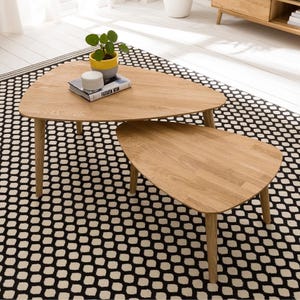 May include: Two light oak coffee tables with rounded triangular tops and slender legs. The tables are nested, with a smaller one beneath a larger one. A plant in a yellow pot and a book are on the larger table. The tables sit on a black and white patterned rug.