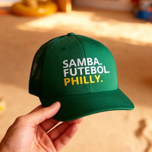 May include: Green baseball cap with the words "SAMBA. FUTEBOL. PHILLY." in white and yellow lettering. The cap has a mesh back and a curved brim. The cap is being held up by a hand.