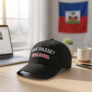 May include: Black baseball cap with the text "SAK PASSE? GOAL PASSE." in white and red lettering. The cap is on a wooden surface, with a laptop and a Haitian flag in the background.