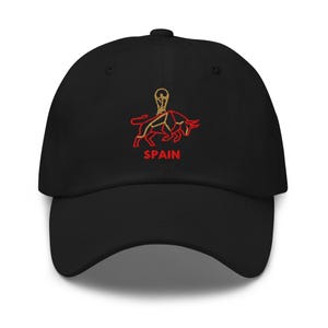 Puede incluir: Gorra de béisbol negra con un diseño bordado de un toro y un trofeo, con la palabra "SPAIN" en rojo. La gorra tiene una visera curva y una correa ajustable.