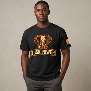 May include: Black t-shirt featuring an orange and tan elephant graphic with the words "Tusk Power" in a rectangular orange frame. The shirt has a small flag on the sleeve.