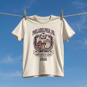 May include: Cream-colored t-shirt with a graphic design featuring the text "PHILADELPHIA, PA" and a soccer ball. The design includes illustrations of city landmarks and the text "KICKOFF IN THE CITY OF BROTHERLY LOVE" and "2026".