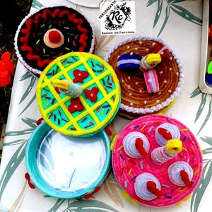 May include: A collection of colorful, handmade, circular containers with decorative, felt-like toppings. The containers are in various colors, including pink, brown, yellow, and teal, with embellishments like sprinkles and flowers. The text "TERIMAKASIH" and "Ranran Collections" are visible.