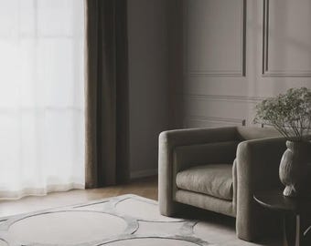 May include: A beige upholstered armchair with rounded armrests and a thick seat cushion, positioned on a large area rug featuring a repeating pattern of interlocking circles in shades of gray and beige, next to a small dark side table with a vase.