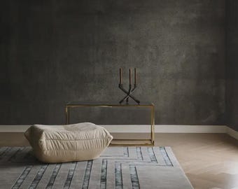 May include: A rectangular area rug featuring a gray surface with a repeating geometric pattern of dark gray zig-zag lines. The rug is positioned on a light brown wooden floor, accompanied by a beige floor cushion, a gold console table, and a candle holder.