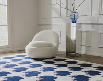 May include: A rectangular area rug featuring a repeating pattern of dark blue geometric shapes with light purple outlines on a cream-colored background. The rug is positioned on a wooden floor near a white rounded chair and a mirrored pedestal.