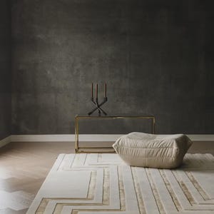 May include: A rectangular area rug featuring a geometric pattern of raised cream-colored lines and recessed gold-toned sections. The rug is placed on a wooden floor next to a beige, soft-textured floor cushion, a gold-framed console table, and a candle holder.