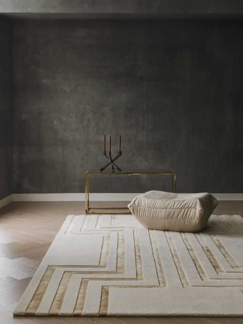 May include: A rectangular area rug featuring a geometric pattern of raised cream-colored lines and recessed gold-toned sections. The rug is placed on a wooden floor next to a beige, soft-textured floor cushion, a gold-framed console table, and a candle holder.