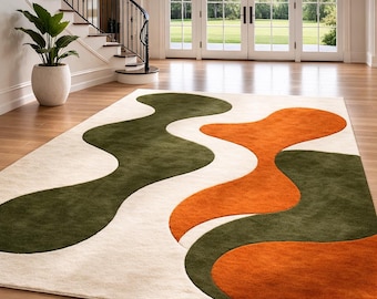 May include: A rectangular area rug with a cream-colored background and abstract wave-like patterns in olive green and orange. The rug is placed on a hardwood floor in a well-lit room with a staircase and large windows.