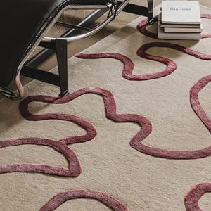May include: Beige area rug with abstract, wavy, burgundy-colored design. A black leather chair with a chrome frame is in the background, along with a stack of books. The rug's design adds a modern touch to the room.