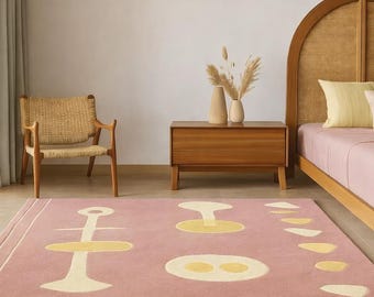 May include: A pink area rug with a geometric design featuring yellow and off-white shapes. The rug is in a bedroom setting, with a wooden nightstand, a woven chair, and a bed with a woven headboard. The rug's design adds a touch of modern art to the room.