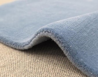 May include: Close-up of a section of a light blue rug with a soft, plush texture. The edge of the rug is visible, showing a neatly stitched border. The rug is slightly curved, revealing its thickness and construction.