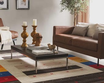 May include: A rectangular area rug featuring a cream-colored center with a dark brown border, accented by small red animal motifs and geometric blocks of blue and yellow at the corners. The rug is positioned beneath a dark coffee table in a living room.